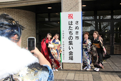 はたちの集いの立て看板の左右でピースする4名の振り袖姿の女性その様子をスマートフォンで撮影する女性